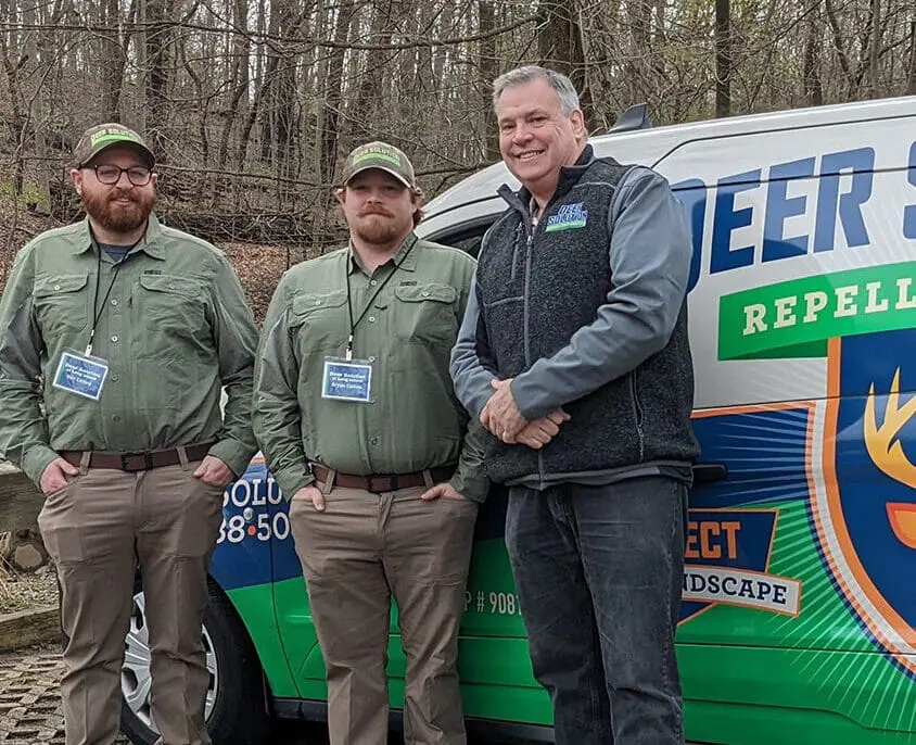 Father and son, Bill and Bryan Collins, new franchisees of Deer Solution, standing in front of a branded Deer Solution van, wearing company uniforms and badges, smiling in an outdoor setting.